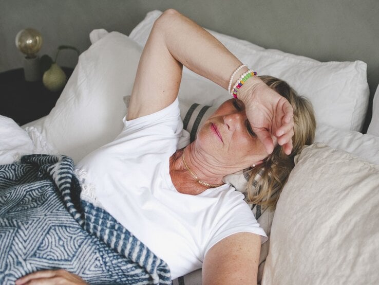 Zu sehen ist eine Frau mittleren Alters im Bett liegend, der sichtlich zu warm ist.