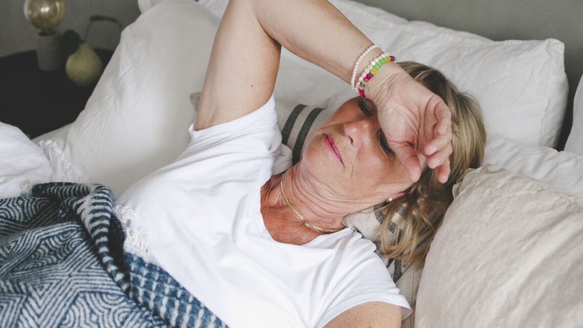 Zu sehen ist eine Frau mittleren Alters im Bett liegend, der sichtlich zu warm ist.