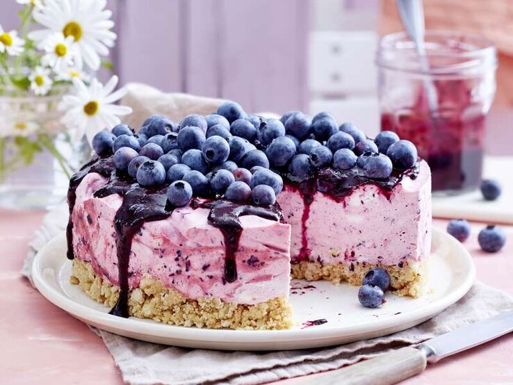 Cremiger Heidelbeer-Cheesecake für deine Gartenparty gefällig? Heidelbeer-Cheesecake mit einem Topping aus frischen Heidelbeeren und einem Boden aus Butterkeksen, serviert auf einem hellen Tisch gedeckt mit rosa Tischdecke.