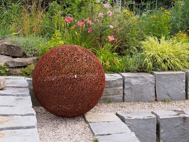 Rostrote Kugelskulptur aus Metall steht dekorativ auf Kies vor einem blühenden Staudenbeet und einer Natursteinmauer.