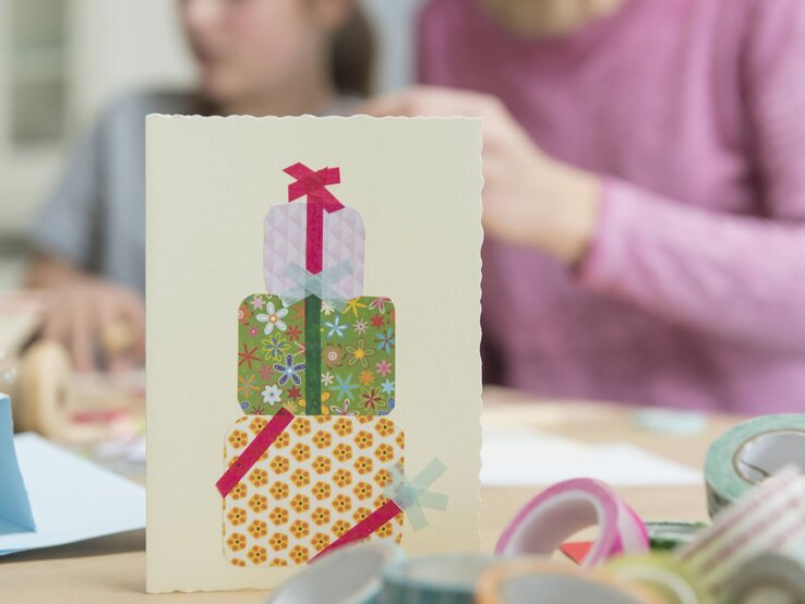Selbstgebastelte Grußkarte mit bunten Geschenkpaketen aus Washi-Tape steht im Vordergrund, zwei Personen basteln im Hintergrund.