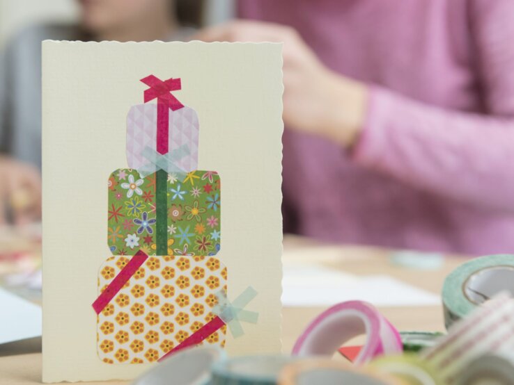 Selbstgebastelte Grußkarte mit bunten Geschenkpaketen aus Washi-Tape steht im Vordergrund, zwei Personen basteln im Hintergrund.
