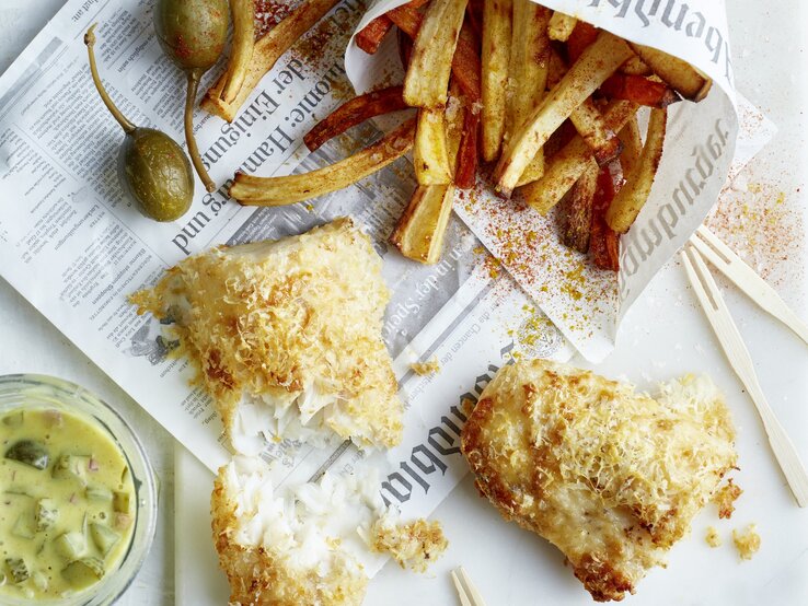 Rustikales Fish-and-Chips-Gericht mit Pommes in Papier, kross gebackenem Fisch, Zitronenspalten und Glas Remoulade.