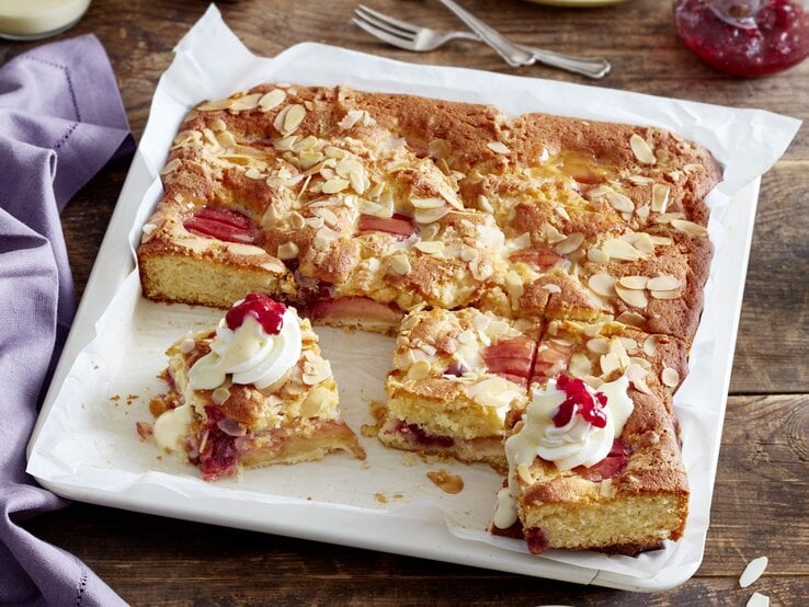 Versunkener Apfel-Sahnekuchen  Saftiger Versunkener Apfel-Sahnekuchen auf einem Backblech, dekoriert mit Sahnetuffs, Preiselbeeren und gehobelten Mandeln, auf einem rustikalen Holztisch.
