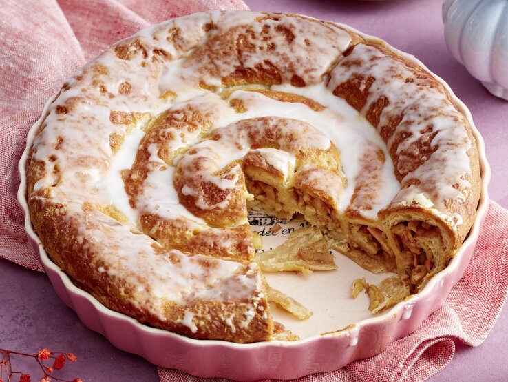Frisch gebackene Croissant-Apfel-Schnecke in einer rosa Backform, überzogen mit glänzendem Zuckerguss und gefüllt mit Zimt und Apfelstückchen, auf einem lila Hintergrund mit rosafarbenem Tuch.