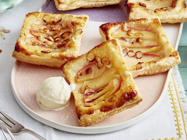 Vier Blätterteigtörtchen mit Apfelscheiben und gehobelten Mandeln liegen auf einem Teller neben einer Kugel Vanilleeis.