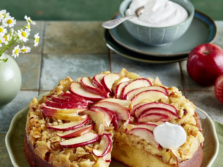Frischer Apfelkuchen mit glänzenden Apfelscheiben und Mandeln auf grünem Teller, dekoriert mit Kamillenstrauß. | © EAT CLUB/Isabell Triemer