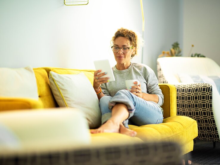 Eine Person mit lockigen Haaren sitzt entspannt auf einem gelben Sofa mit Kissen, hält ein Tablet und eine Tasse in einem hellen Raum.