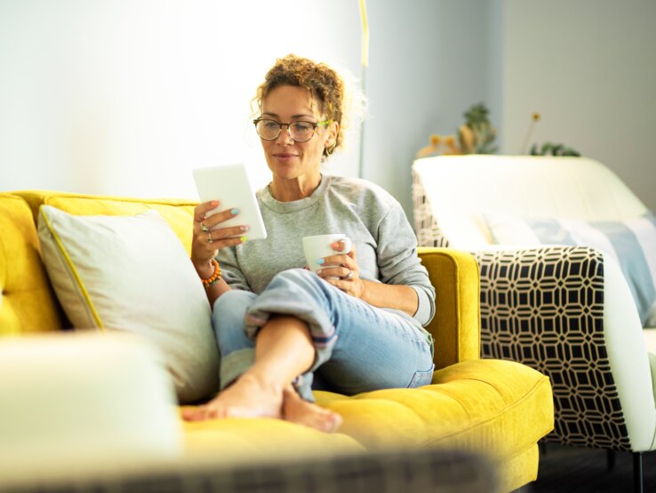 Eine Person mit lockigen Haaren sitzt entspannt auf einem gelben Sofa mit Kissen, hält ein Tablet und eine Tasse in einem hellen Raum.