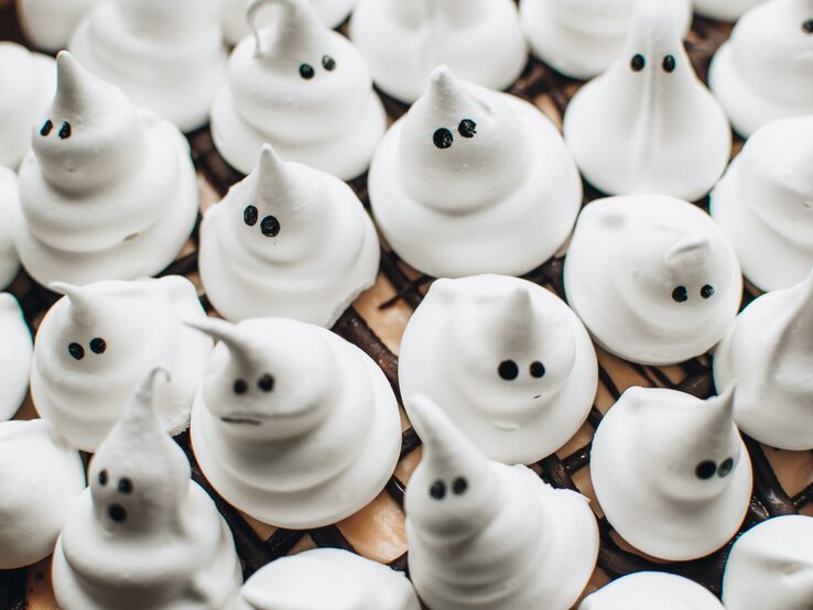 Gruseliger Naschspaß: Unsere Geisterbaiser sind perfekt für deine Halloween-Party! Kleine weiße Geister aus Baiser mit lustigen Gesichtern sitzen eng nebeneinander auf dunklem Gittermuster.