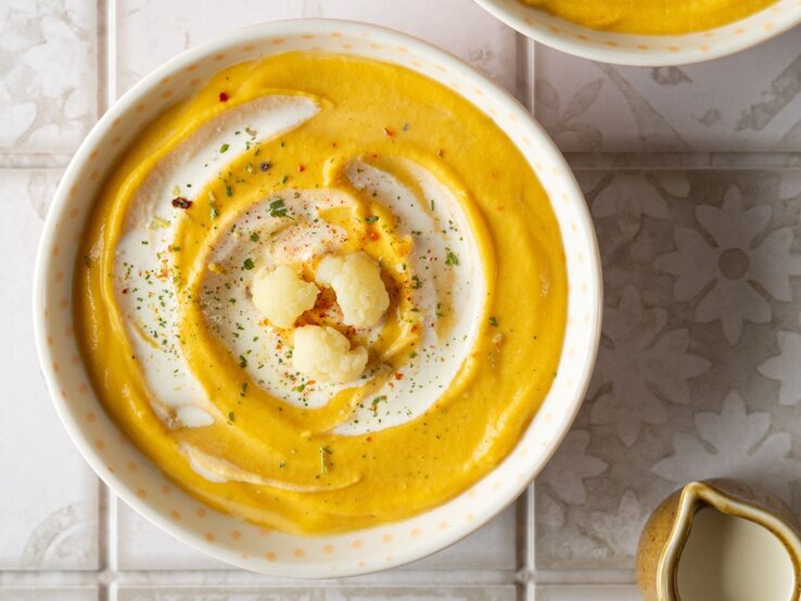 Zwei Schüsseln mit cremiger Kürbis-Blumenkohl-Suppe, dekoriert mit einem Swirl aus Joghurt und gerösteten Blumenkohlröschen, auf einer hellen, gemusterten Fliesenoberfläche. Neben den Schüsseln steht eine mit Suppe gefüllte Pfanne und eine kleine Kanne, umgeben von beigen Stoffservietten.