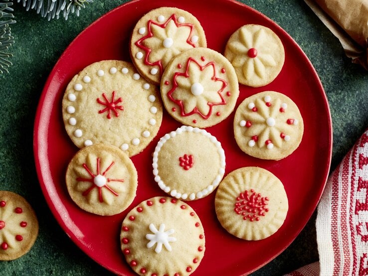 Elf liebevoll verzierte Vanille-Plätzchen mit filigranen Mustern in Rot und Weiß auf rotem Teller auf grünem Untergrund.