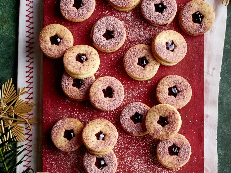 Brombeer-Marzipan-Kekse mit dunkler Marmelade und rosafarbenem Zucker auf rustikalem Brett, daneben Sieb und Deko.