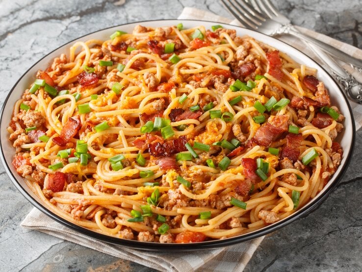 Cowboy Spaghetti mit Hackfleisch, Speck, Tomatenstücken und Frühlingszwiebeln auf einem Teller mit Besteck auf Marmorplatte.