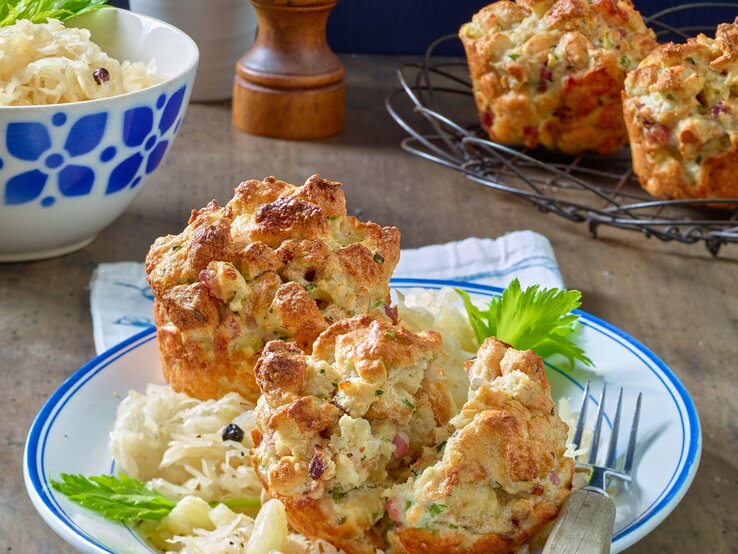 Zwei Speck-Semmelknödel-Muffins mit dem Ananas-Sauerkraut auf einem Teller von vorne fotografiert, im Hintergrund weitere Muffins und eine Schale mit Sauerkraut.