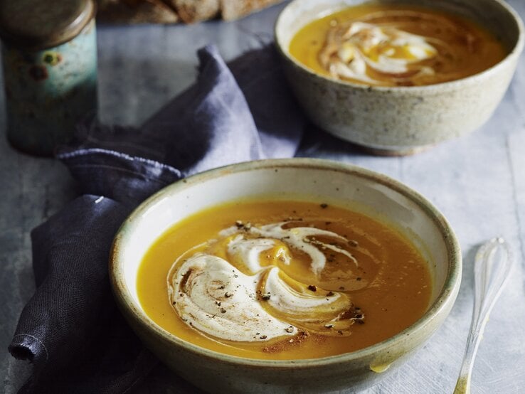 Zwei Schalen Kürbissuppe mit Sahne und Zimt stehen auf grauem Stein, daneben ein Löffel, Leinentuch und frisches Sauerteigbrot.