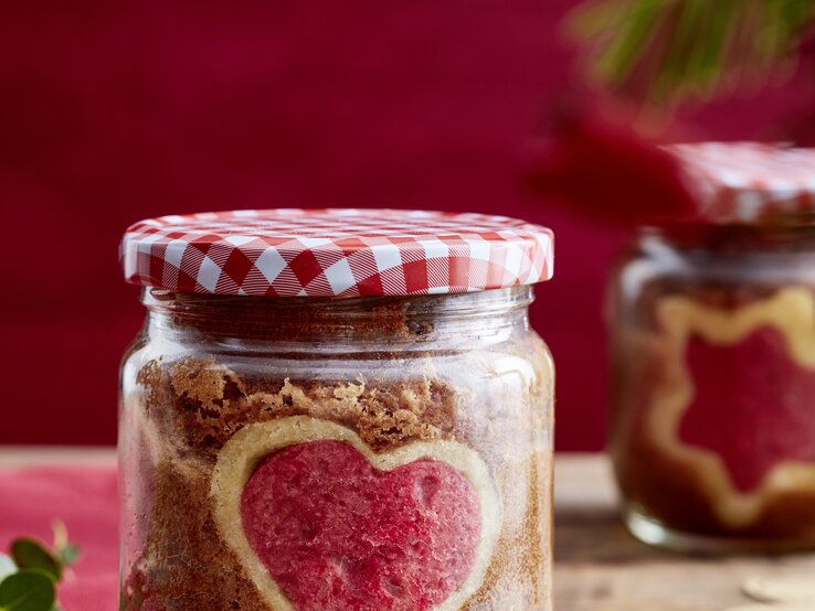Selbstgebackener Kuchen mit rotem Herz in einem Schraubglas mit Karodeckel, daneben ein Geschenkanhänger auf Holz.