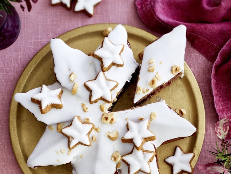 Weihnachtlicher Zimtsternkuchen mit glänzendem Zuckerguss, Zimtsternen und Haselnüssen auf edler Goldplatte, vor rosa Tischwäsche und Deko.