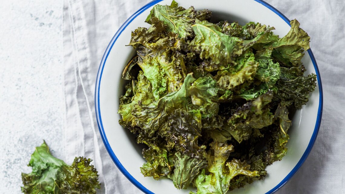 Draufsicht auf eine Schüssel mit knusprigen Grünkohl-Chips, daneben ein einzelnes Blatt auf einem hellen Untergrund, arrangiert auf einem Tuch.