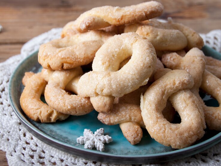 Auf einem türkisfarbenen Teller liegen goldbraune, gezuckerte Berlinerkranser auf einem weißen Häkeldeckchen.