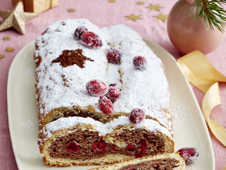 Festlicher Spekulatius-Stollen mit Puderzucker und kandierten Cranberrys auf Platte, daneben goldene Deko auf rosa Stofftischdecke.