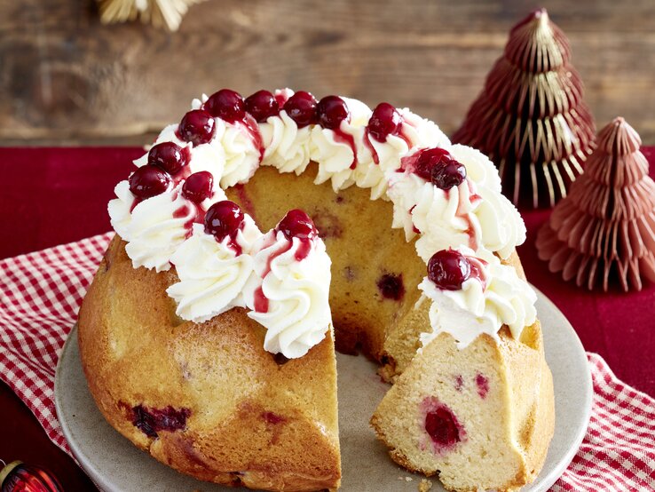 Aufgeschnittener Glühwein-Kirsch-Hupf mit Sahnehaube und Fruchtsoße steht festlich dekoriert auf einem grauen Teller mit Stoffserviette.