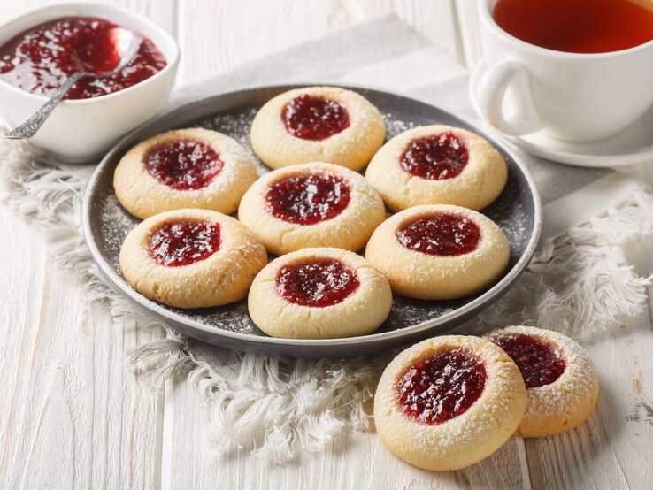 Frisch gebackene Marmeladen-Kekse mit Zuckerschnee serviert auf Leinentuch, daneben dampfender Tee in weißer Tasse.