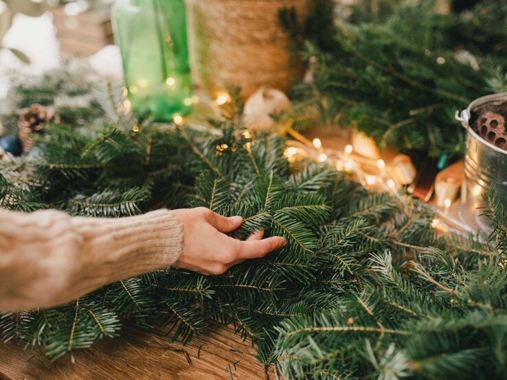 Auf einem Holztisch arrangiert eine Person mit hellem Pullover duftende Tannenzweige zwischen warmen Lichterketten.