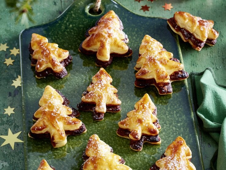 Schräge Draufsicht auf ein Blech mit frisch gebackenen Nougat-Tannenbäume vor einer weihnachtlichen Kulisse.