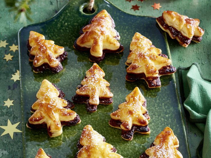 Schräge Draufsicht auf ein Blech mit frisch gebackenen Nougat-Tannenbäume vor einer weihnachtlichen Kulisse.