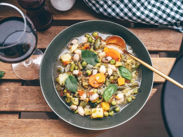 Draufsicht auf einen tiefen, dunklen Teller mit einer Minestrone aus viel Gemüse, der auf einem Tisch neben einem Weinglas steht.