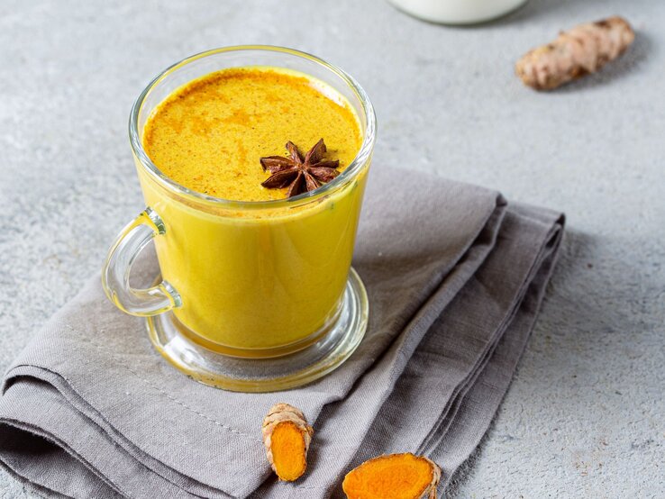Ein Glas Goldene Milch, garniert mit einem Sternanis, steht auf einer grauen Stoffserviette. Daneben liegen frische Kurkuma- und Ingwerstücke.
