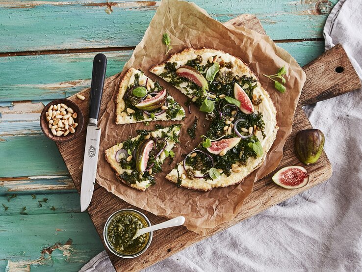 Draufsicht auf die Low-Carb-Pizza mit Grünkohlpest, Feigen und Pinienkernen, die auf einem Holzbrett auf einem türkisen Untergrund liegt.