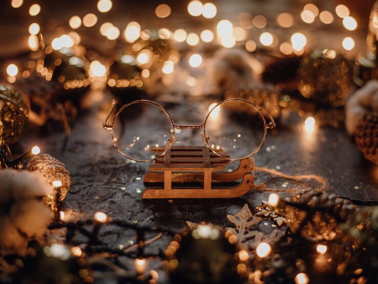 Runde goldene Brille von Harry Potter liegt auf einem kleinen Holzschlitten, umgeben von Tannenzapfen und warmen Lichterketten.