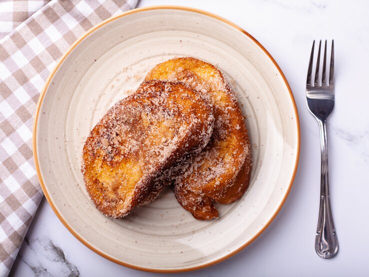 Draufsicht auf einen Teller mit zwei Scheiben Arme Ritter, die mit Zimt und Zucker bestäubt sind.