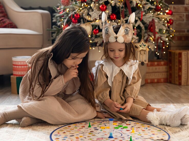 Zwei Mädchen in beigefarbenen Kleidern spielen konzentriert ein Brettspiel vor festlich geschmücktem Weihnachtsbaum.