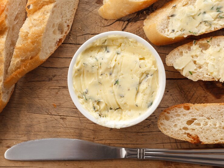 Butter mit Zitronenverbene in einer kleinen runden Form / Baguette ganz und aufgeschnitten / Streichmesser aus Metall / Untergrund grobes Holz