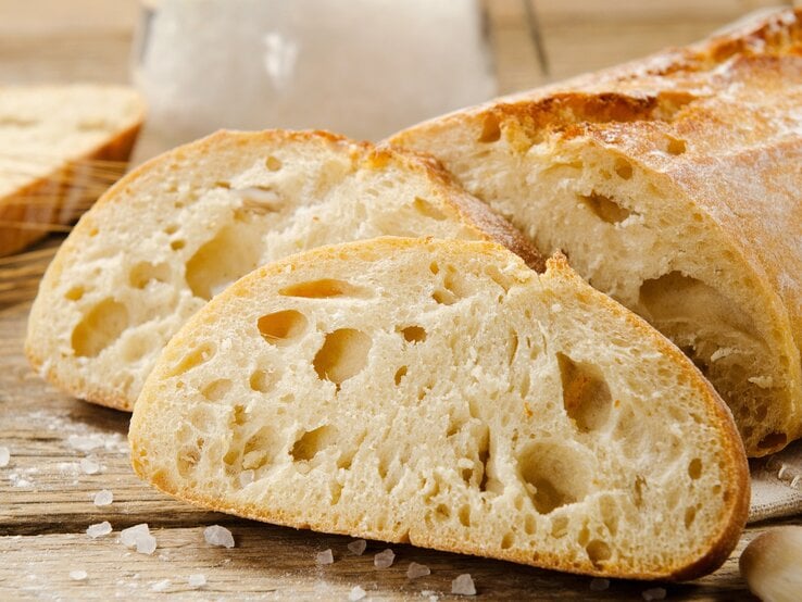 Ciabatta: zwei Scheinen und  der angeschnittene Brotlaib / einige Krümel Meersalz / Untergrund grobes Holt / Frontalsicht