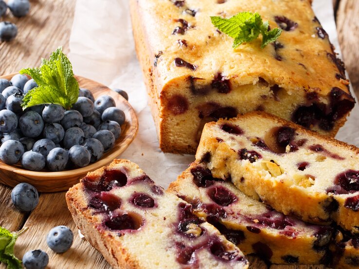 Zitronenkuchen mit Heidelbeeren / angeschnitten und einzelne Scheiben / runde Holzschale mit frischen Meidelbeeren / Minzblätter / Untergrund Holz / leicht schräge Drauf- und Frontalsicht