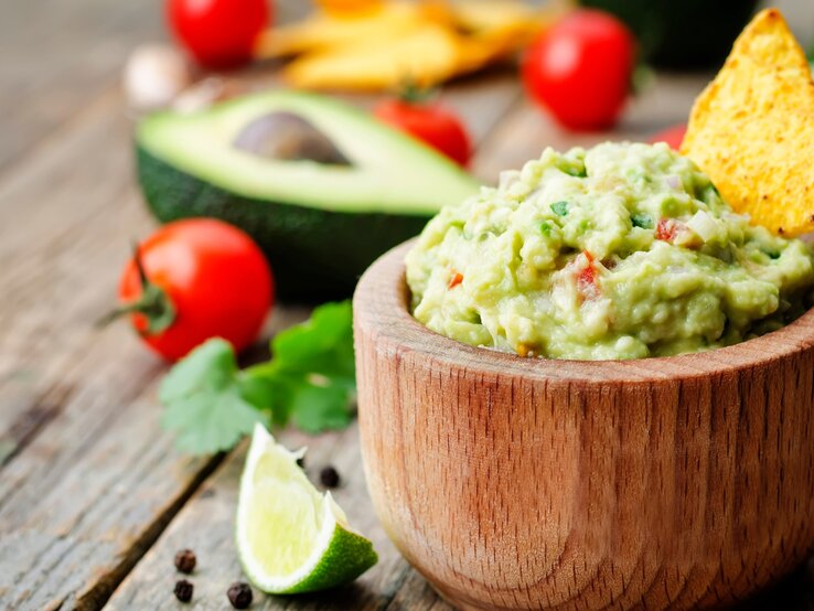 Guacamole in runder Holzschüssel / Nacho-Chip in der Guacamole / einzelne Tomaten, halbierte Avocado / Korianderblatt / Pfefferkörner / Untergrund grobes Holz
