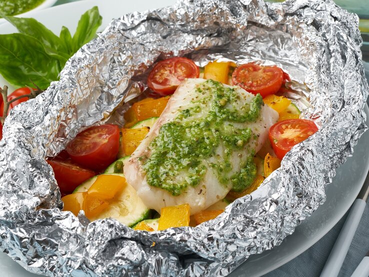 Fischfilet-Päckchen aus dem Ofen in Pesto bestrichen / In Alufolie gewickelt mit Tomaten, Paprika und Zucchini / Draufsicht schräg