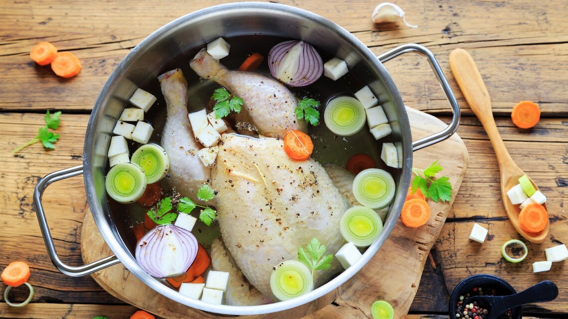 Hühnerbrühe im Topf / ganzen Suppenhuhn, klein geschnittenes Gemüse / Unterlagen Holzbrett auf Holzboden / frischer Pfeffer in Mörser / Holzlöffel / frisches geschnittenes Gemüse - Möhren, Knoblauch, Sellerie, Petersilie