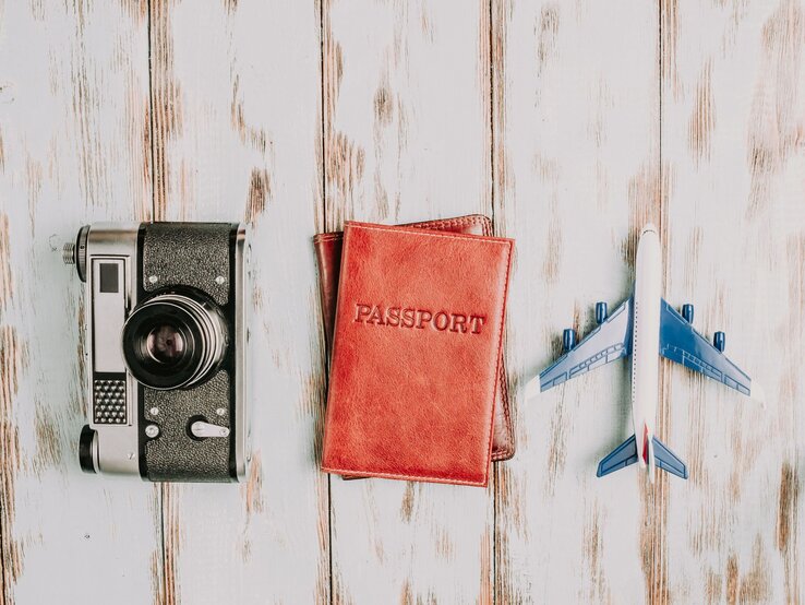 Es sind eine rote Geldbörse, ein Fotoapparat und ein Spielzeugflugzeug liegend auf weiß-grauem Untergrund zu sehen.