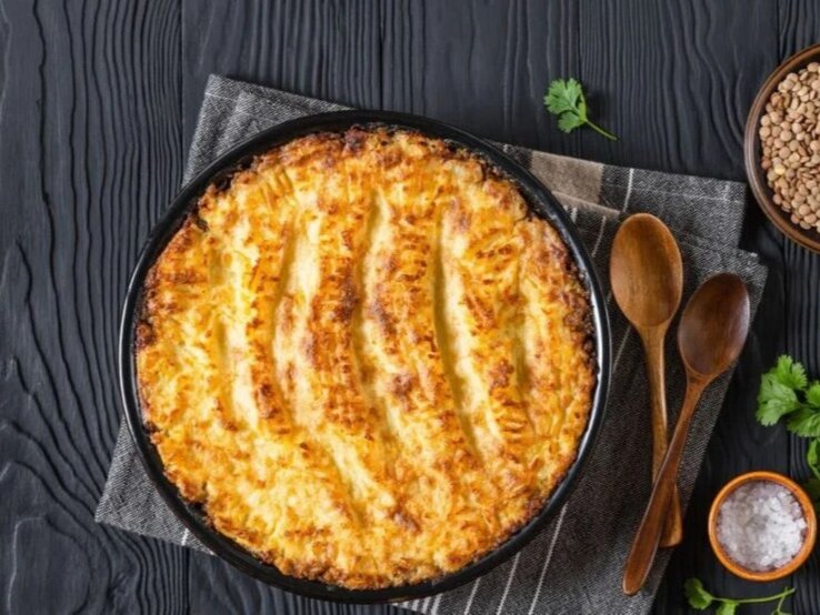 Veganer Linsen-Gemüse-Auflauf in runder Auflaufform auf dunklem Holztisch mit Linsen und Kräutern.