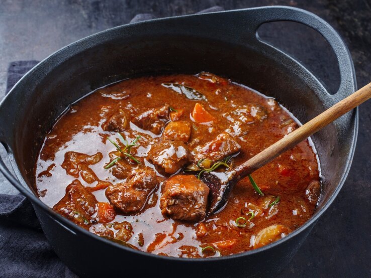 Ungarisches Gulasch wie bei Oma / Fleischstücke, Rosmarin, Möhrenstück / Holzlöffel im Topf / Draufsicht, schräg