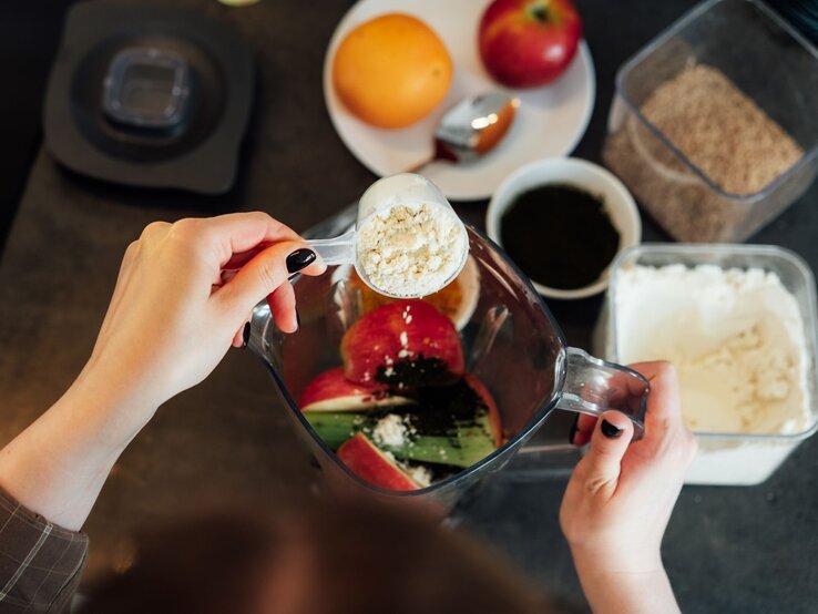 Zwei Hände füllen gesundes Obst und Gemüse sowie Kollagenpulver in einen Mixer.