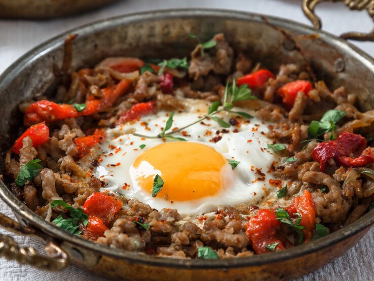 Kiymali Yemek mit Spiegelei und frischen Kräutern in einer runden Pfanne aus Metall / Gabel / Untergrund Holz