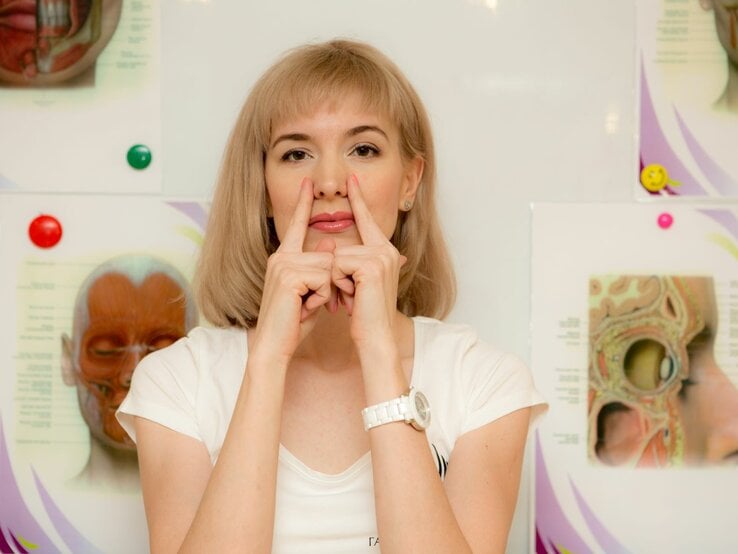 Eine Frau führt Gesichtsyoga-Übungen durch, indem sie mit den Zeigefingern ihre Nase nach oben drückt, vor anatomischen Postern des menschlichen Gesichts.