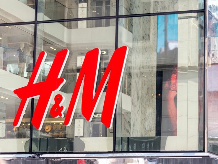 Das Bild zeigt das große, leuchtend rote Logo von H&M an der Fassade eines Geschäfts. Durch die großen Fenster hinter dem Logo sind Mannequins und Teile der Inneneinrichtung des Ladens zu erkennen.